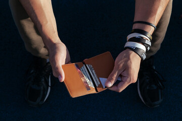 a young man in sportswear is holding a small comfortable leather wallet with dollar bills in his hands. top view. Horizontal orientation, copy space, close-up, no face - obrazy, fototapety, plakaty