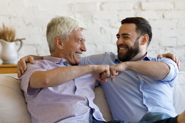Happy older 50s man his young adult son sit on sofa at home smiling make fist bump gesture, showing...