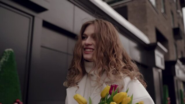 Nice girl in white coat with bouquet of tulips is walking down the street. Spring and tulips. Smile. High quality 4k footage