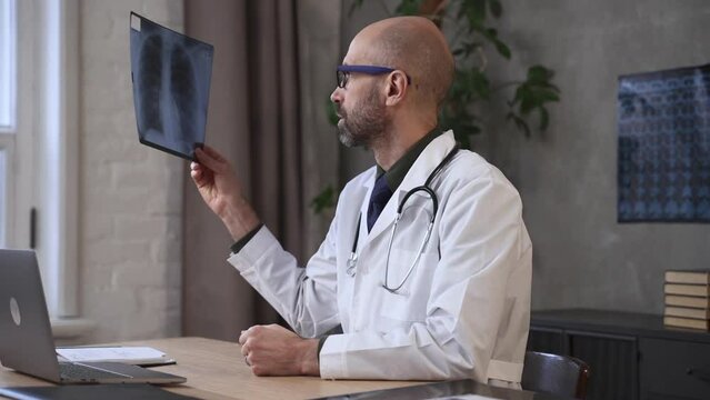 Male doctor looking at x-ray and sitting at table with device in clinic office spbas. Close view of caucasian bearded man holds picture and examines carefully, makes diagnosis and sits at desk with