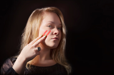 A young girl draws two stripes on her cheek with her fingers using lipstick War paint