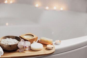 Fototapeta na wymiar Preparation for hotel spa treatment or home bath procedure. White washbasin in bathroom with accessories on tray. Burning candles, washcloth, soap, foot brush, bottle with sea salt, orchid flower