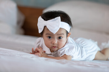 Cute adorable baby girl lying on white bed looking at camera cheerful and comfortable.Infant asian baby wear white dress and headband on bed.Infant baby concept