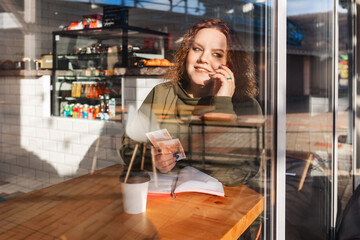 Portrait of a fatty smiling woman sitting in a cafe and calling at the cellphone. View through the cafe window, double exposure. The concept of business and freelancing