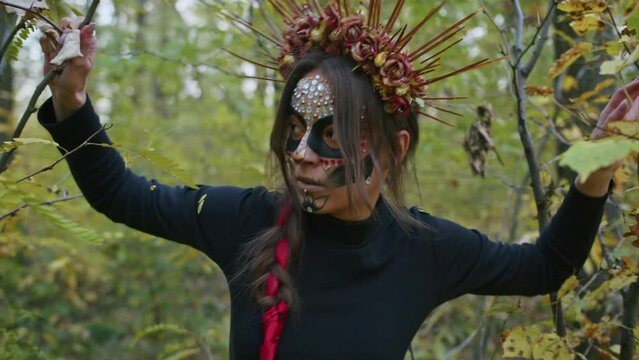 A young woman with Santa Muerte make-up dressed in a black dress of death walks against the backdrop of autumn leaves in the forest during sunset. Day of the Dead or Halloween concept.