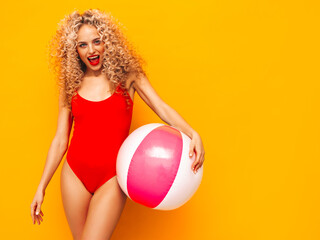Fototapeta na wymiar Young beautiful smiling woman posing near yellow wall in studio.Sexy model in red swimwear bathing suit.Positive female with curls hairstyle. Holding penny inflatable ball.Happy and cheerful