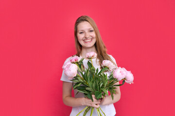 International women's day. Extremely happy woman in white t-shirt sniffing bouquet of spring flowers pink peonies that she holds in her hands on a bright pink background in the studio