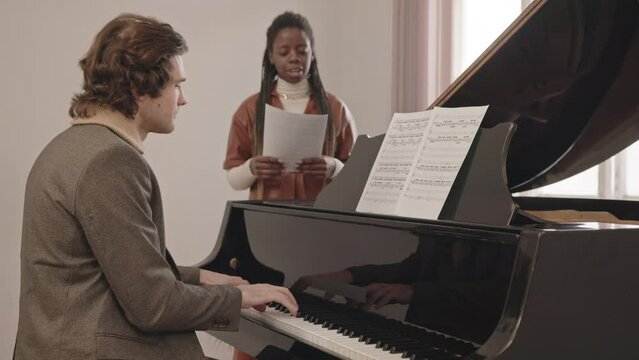 Medium slowmo of young interracial music duo having rehearsal indoors. Caucasian man playing grand piano while African-American woman with lyrics in hands singing