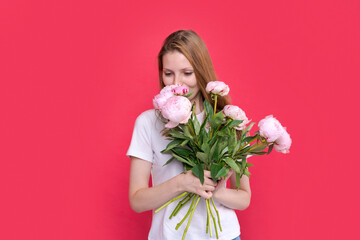 Portrait cheerful young lady with romantic spring bouquet of pink peonies on a bright pink studio background. Gorgeous girl enjoys women's day celebration and receives flowers as a special gift