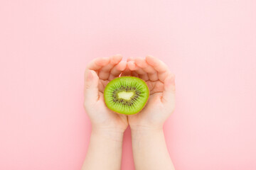 Baby girl hands holding half of fresh green kiwi on light pink table background. Pastel color. Closeup. Point of view shot. Top down view.