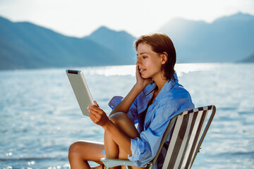 Smiling woman sitting on deck chair by the sea using tablet on a sunny day