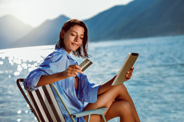 Happy woman with digital tablet and credit card buying online sitting near the sea.