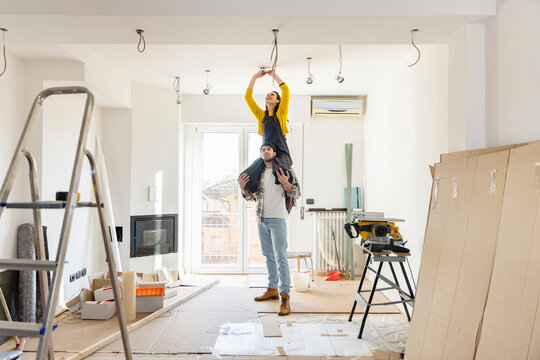 Young man carrying girlfriend on shoulders installing light bulb in living room