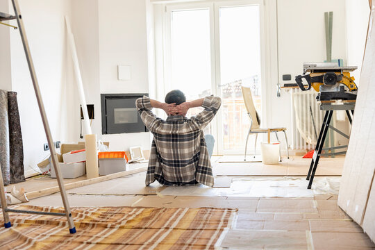 Young man with hands behind head looking through window of living room being renovated