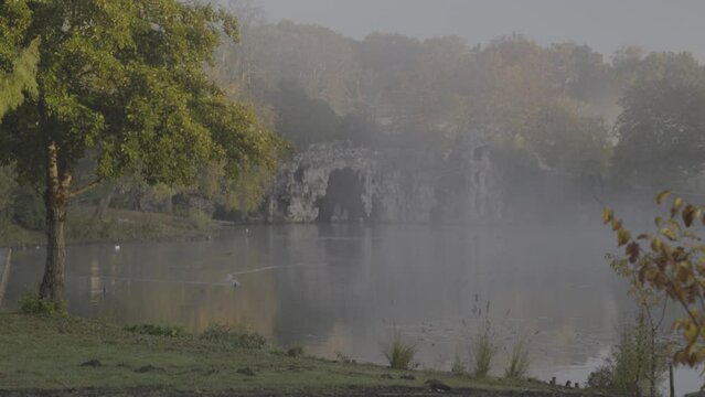 Grotto cave at misty Majolan Park lake in Blanquefort France with ducks and geese swimming, Locked shot
