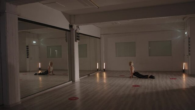 Blonde lady stretching in front of huge mirror in empty dance studio. Wide long shot, slow motion
