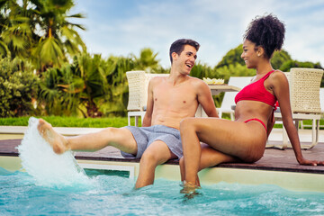 Biracial couple of young people in love having fun sitting by the pool chatting and splashing the water with their feet