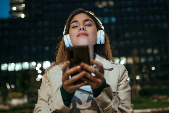 Woman With Eyes Closed Holding Mobile Phone Listening Music Through Headphones At Night