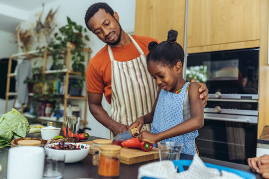 Girl cutting pointed pepper by father in kitchen at home
