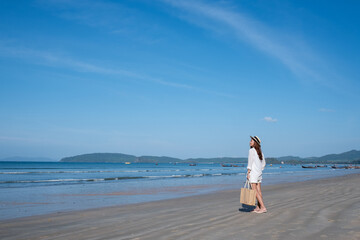Fototapeta na wymiar Portrait image of a beautiful young asian woman with hat and bag strolling on the beach with the sea and blue sky background