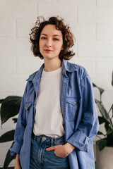 Caucasian young woman with short curly hair standing in front of white wall and green leaves looking at camera