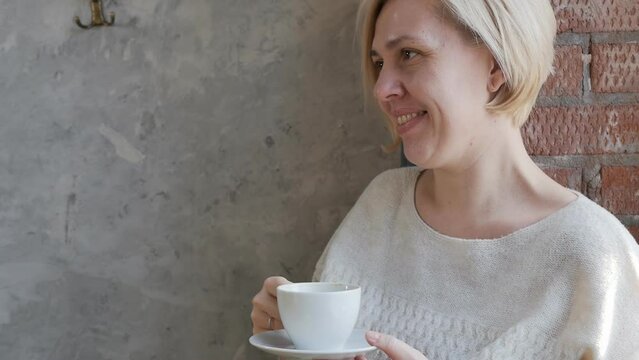 Portrait. A young European blonde with a caret holds a cup of tea and listens attentively to an interlocutor in a cafe at a table. Against the background of a gray wall and brickwork