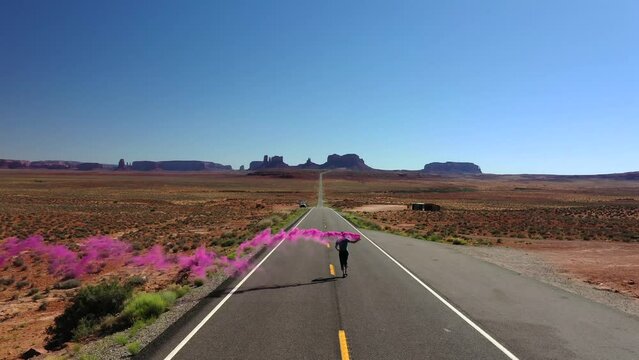 Man Running On The Highway In Utah Holding Color Smoke Signal. Monument Valley In Distant Background. wide aerial