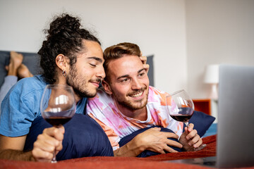 young male gay couple watching movie in bed on laptop while drinking red wine, bedtime holiday...