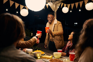 Black male standing, cheers with with group of diverse friends celebrating at an outdoor party . Trendy, young adults