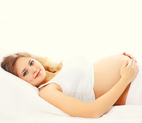 Fototapeta premium Portrait of smiling young pregnant young woman lying on the bed at home in white room
