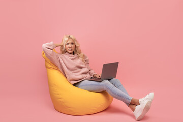 Troubled lady sitting on beanbag chair with laptop, making mistake in online project, pink background, copy space