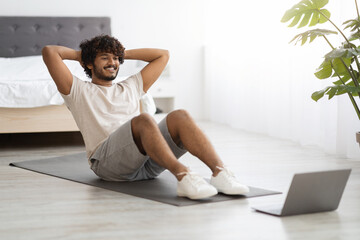 Active hindu guy exercising at home, using laptop