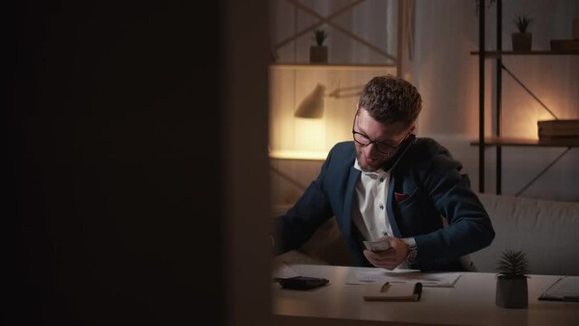 Business tasks. Busy man. Financial accounting. Elegant rich guy counting cash money talking mobile phone sitting desk in dark light room interior.