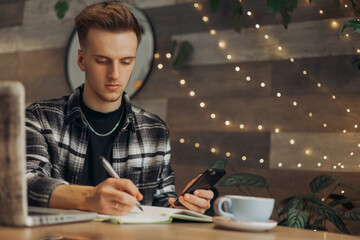 Pensive young male freelancer taking notes in notepad and working in cafe while using smartphone 