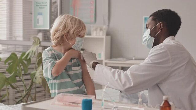 Medium slowmo shot of 7-year-old Caucasian boy getting vaccinated by African-American young male doctor in modern clinic