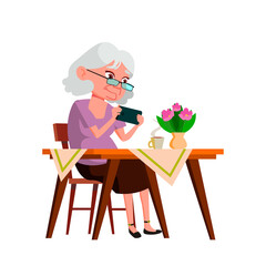 Old Woman Grandma Playing On Smartphone Vector. Elderly Lady Sitting At Table, Drinking Hot Drink And Play On Smartphone Digital Device. Character Use Phone At Desk Flat Cartoon Illustration