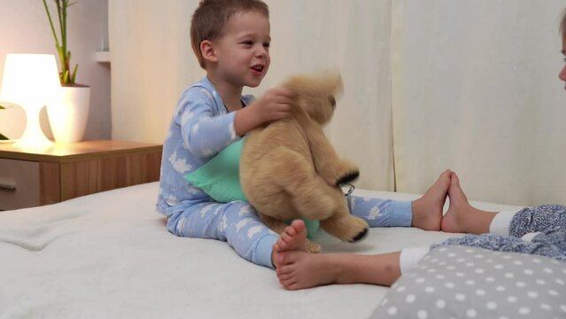 Two Smiling Preschool Toddler Children In Pajamas Playing With Teddy Bear on Bed. Siblings Little Twins Boy and Girl Have Fun. Happy Kids On Quarantine At Home. Friendship, Family, Education Concept