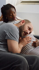 Multi ethnic parents talking about baby and parenting while sitting together on couch. Caucasian father of child touching belly of african american wife at home. Interracial couple