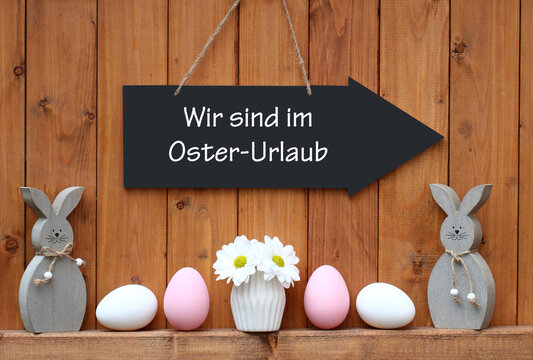 Schild mit dem wir sind in Osterurlaub vor einer Holzwand mit Osterdekoration.