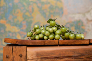 A bunch of unripe green cherry tomatoes lying on an old wooden table, and a multi-colored wall in the background.