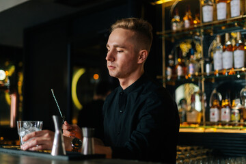 Professional young barman standing behind bar counter and holding stirring spoon and glass filled with ice cubes, preparing to make cocktail, on background of shelves with alcoholic drinks.