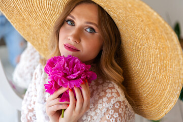 Close-up portrait of a beautiful woman in a straw hat, looking at the camera. Woman with makeup and red lips, red peony in her hands. Blonde with curled hair