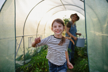 Portrait of happy little farmer girl with grandmother in greenhouse, looking at camera.