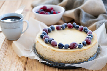 Vanilla cheesecake decorated with blueberry and cranberry on parchment with coffee cup and forks on background. Festive dessert pastry.