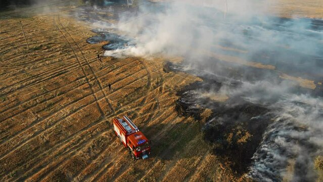 Fire truck at hearth of dry stubble flame and firefighters extinguish huge burning of field with water from hoses. Emergency case for danger mission and rescue nature saving concept