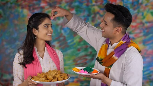 Two youngsters celebrating the festival of Holi by putting Gulal Tilak on the forehead. A cheerful girl wearing a white Kurti holding a plate of traditional Gujia - an Indian festival of colors  a ...