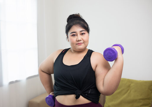 Attractive asian young fitness woman plus size lifting dumbbell weights workout at home in living room. Female training and exercise wearing sport wear fit body. Happy and is proud of herself.