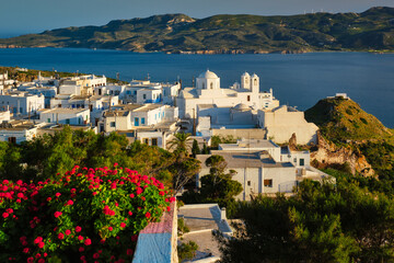 Picturesque scenic view of Greek town Plaka on Milos island over red geranium flowers and Orthodox greek church. Plaka village, Milos island, Greece. Focus on buildings