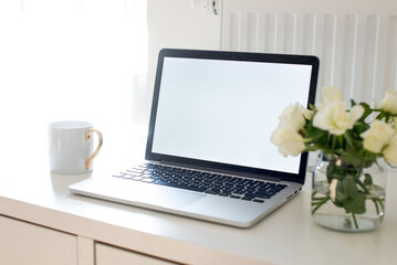 Fototapeta premium Laptop with white blank screen on white work table. White interior home office concept. Female office desk workspace with white roses and cup of coffee. Close up on screen laptop. Mock up laptop