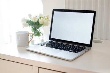 Laptop with white blank screen on white work table. White interior home office concept. Female office desk workspace with white roses and cup of coffee. Close up on screen laptop. Mock up laptop